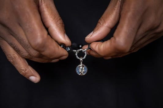 "Hands presenting a Compass-Jewelry black beaded bracelet with a blue sapphire pendant, against a dark backdrop."